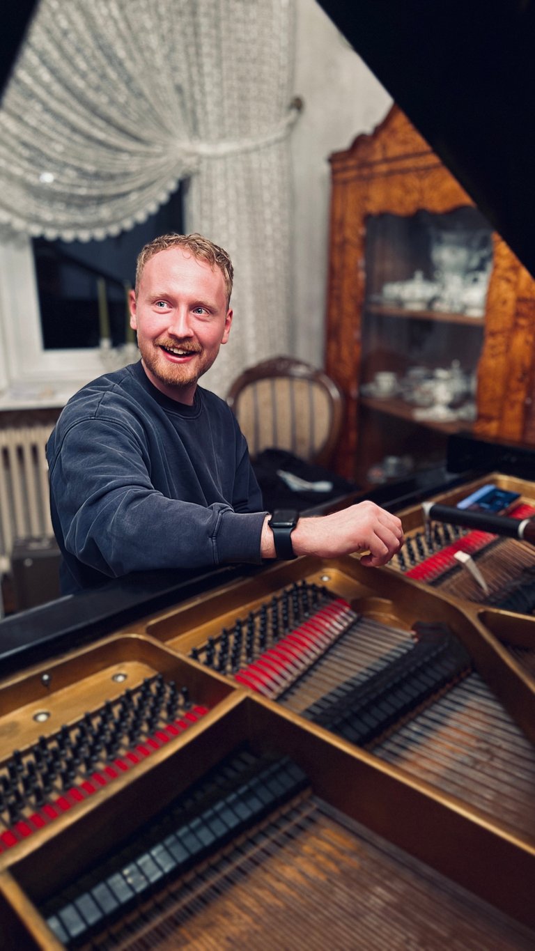 Mann lächelt an einem Flügel in einem elegant eingerichteten Raum mit Samtvorhängen und Holzmöbeln